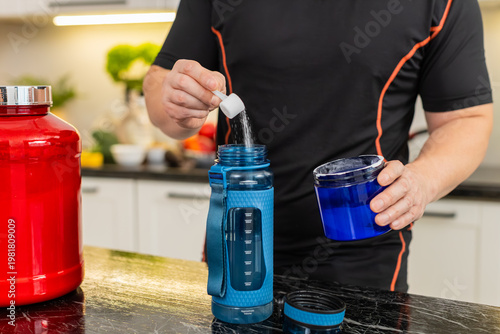 Man adds supplement powder into shaker bottle in kitchen preparing isotonic or creatine drink. Shaking bottle mixes powder with water for workout hydration muscle recovery energy fitness routine gym.