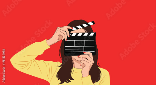 Young woman wearing a yellow sweater holding a movie clapperboard in front of her face against a bright red background.