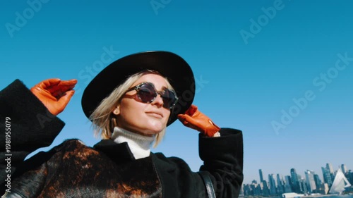 Modern elegant blonde woman in black hat, sunglasses and coat. Low angle view. Blue clear sky and Manhattan skyline in the backdrop.