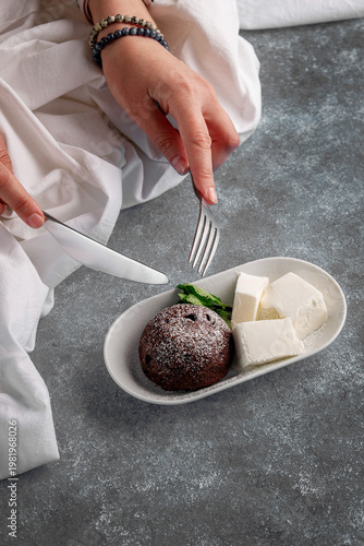 Female hands holding fork and knife over chocolate fondant dusted with powdered sugar, served with vanilla ice cream in elegant dessert presentation