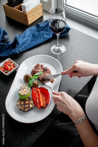 Woman cutting grilled steak served with roasted pepper, zucchini, eggplant, mushrooms, fresh basil, tomato salsa and a glass of red wine
