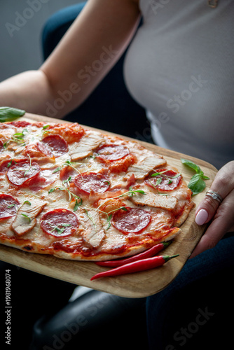 Woman holding a wooden board with baked pizza topped with salami, pork slices, melted cheese, basil and red chili peppers