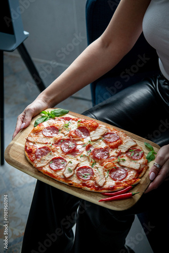 Woman holding a wooden board with homemade meat pizza topped with salami, pork slices, melted cheese, basil and chili peppers