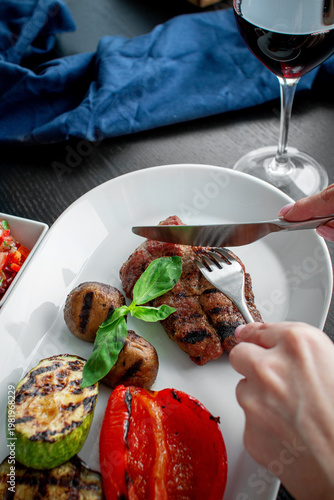 Woman slicing juicy grilled steak served with roasted pepper, zucchini, mushrooms, basil and red wine in elegant restaurant dining presentation