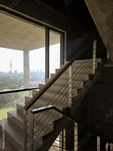 Minimalist concrete staircase design with stainless steel railing and large glass window, modern office or apartment interior with scenic city skyline view, industrial architecture with natural light.