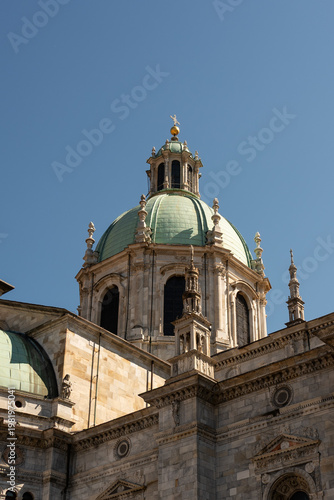 A detailed perspective of the green-domed Como Cathedral against a clear, deep blue sky, showcasing its ornate marble facade and intricate architectural spires.

