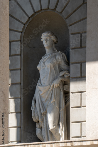 A standing marble statue of a draped female figure within a rusticated stone niche.

