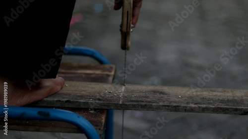 A person cutting a wooden board using a manual hand saw on top of a wooden chair. The scene shows practical carpentry work and tool usage in a simple environment.