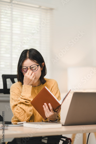 Young Asian woman feeling eye strain and stress while working at home office desk with laptop