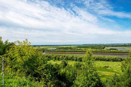 A river flows through a grassy green plain, and hills are visible in the background. The sky is overcast, giving the scene a serene atmosphere.