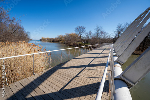 Billede på lærred Curved pedestrian bridge with wooden walkway overlooking calm water and natural shoreline vegetation under a clear blue sky