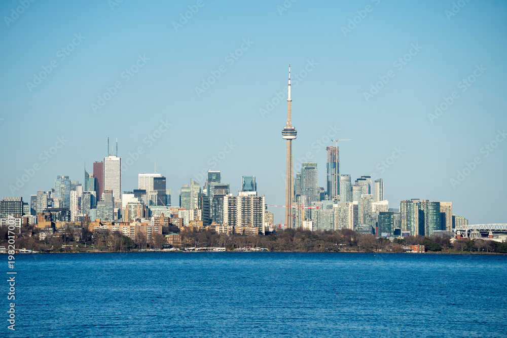 Fototapeta premium Panoramic view of the Toronto skyline featuring the CN Tower, seen from across Lake Ontario on a clear day. The cityscape showcases modern high-rise buildings and waterfront architecture with calm blu