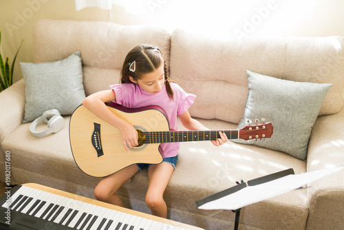 Fotografi Young child practicing chords on a guitar while sitting on a sofa, cultivating h