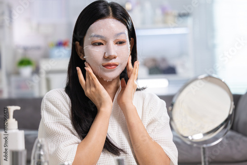 Young Asian woman applying facial sheet mask touching face feeling relaxed peaceful doing skincare routine at home sitting in front of mirror preparing skin treatment for beauty and health