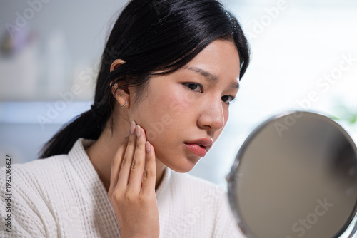 Young asian woman looking into round mirror touching face skin with hand showing worry about cheek acne problem during morning beauty care routine wearing white bathrobe indoors