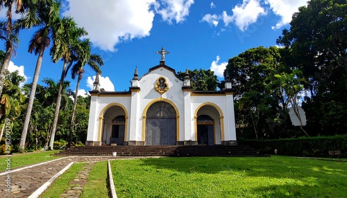 Small white chapel with arched doors and statue, nestled among green trees under a bright, cloudy blue sky