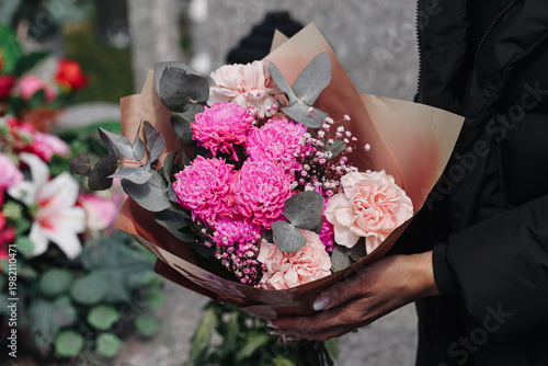 Bouquet of pink flowers held at cemetery during memorial visit. Floral arrangement symbolizing remembrance and respect. Close up of fresh blossoms in hands. Funeral tribute concept outdoors.