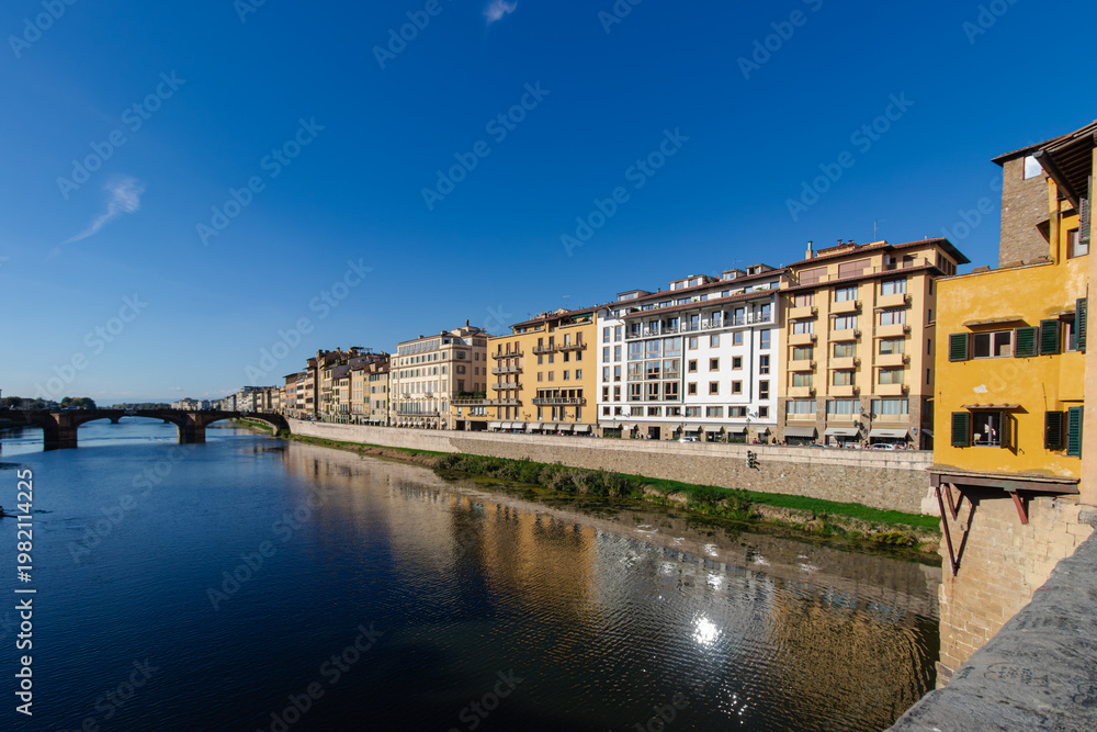 Fototapeta premium Historic Buildings Along Arno River in Florence, Italy with Bridge and Clear Blue Sky