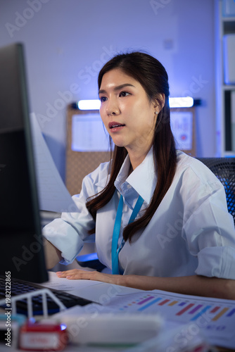 Asian businesswoman working late overtime night modern office typing computer focused dedicated professional looking monitor reviewing data chart graph paperwork desk vertical