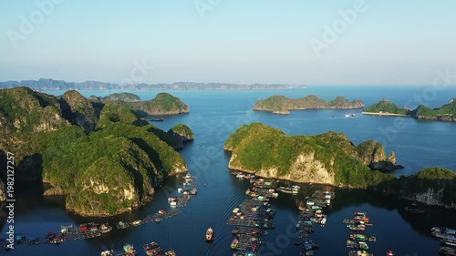 Aerial view of a floating village surrounded by lush limestone islands in Ha Long Bay, Vietnam. Calm blue waters and scenic landscape create a tranquil atmosphere.