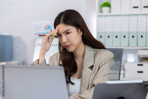 Stressed young asian woman touching head having bad headache from overwork at her office desk looking at laptop screen frustrated and tired from difficult business problem