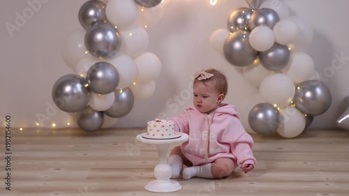 Caucasian baby girl, 1 year old, front view, in pink hoodie with bow, sitting on wooden floor, eating cake, balloons and Happy Birthday sign behind. Concept birthday, childhood, party, celebration.