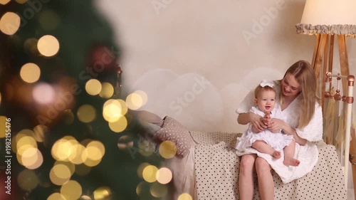 Caucasian young mother in white dress plays with baby girl on bed, front view. Cozy room with soft light, white wall, lamp, Christmas tree blur. Concepts: motherhood, bonding, holidays, love.