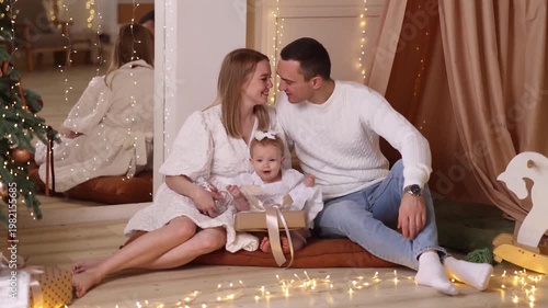 Caucasian parents in white clothes with toddler girl sitting on rug, playing with basket near decorated tree, white wall, wooden floor. Front view. Christmas, family, love, holiday concept.