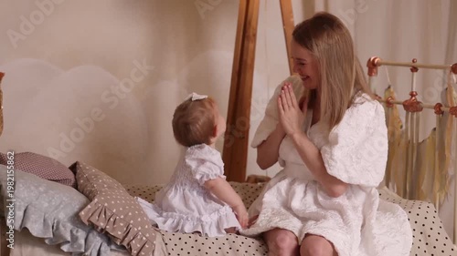 Caucasian young mother in white dress plays with baby girl on bed, front view. Cozy room with soft light, white wall, lamp, Christmas tree blur. Concepts: motherhood, bonding, holidays, love.