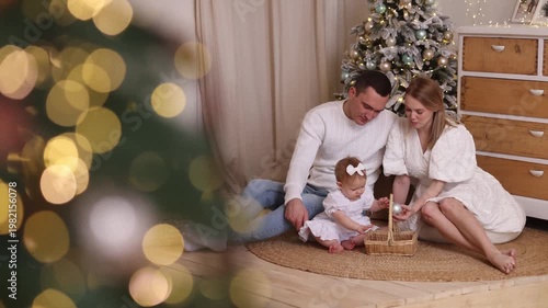 Caucasian parents in white clothes with toddler girl sitting on rug, playing with basket near decorated tree, white wall, wooden floor. Front view. Christmas, family, love, holiday concept.