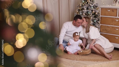 Caucasian parents in white clothes with toddler girl sitting on rug, playing with basket near decorated tree, white wall, wooden floor. Front view. Christmas, family, love, holiday concept.