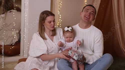 Caucasian parents in white clothes with toddler girl sitting on rug, playing with basket near decorated tree, white wall, wooden floor. Front view. Christmas, family, love, holiday concept.