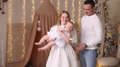 Young Caucasian parents in white clothes hold baby girl in white dress with bow. Front view. Decorated kids room with fairy lights, Christmas tree, beige tent. Concepts: family, holiday, warmth, joy.