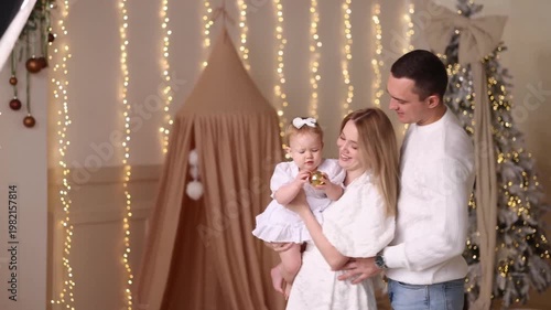 Young Caucasian parents in white clothes hold baby girl in white dress with bow. Front view. Decorated kids room with fairy lights, Christmas tree, beige tent. Concepts: family, holiday, warmth, joy.