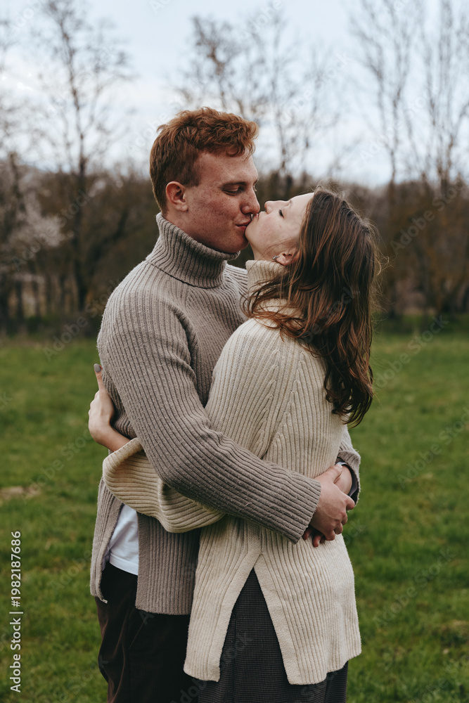 Fototapeta premium Romantic couple embraces and kisses outdoors in knit sweaters, candid lifestyle shot in a park, warm natural light, affectionate moment, serene field setting, intimate connection.