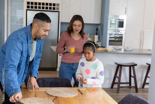 Indian family preparing breakfast in home kitchen, father placing placemat, child inspecting shaker