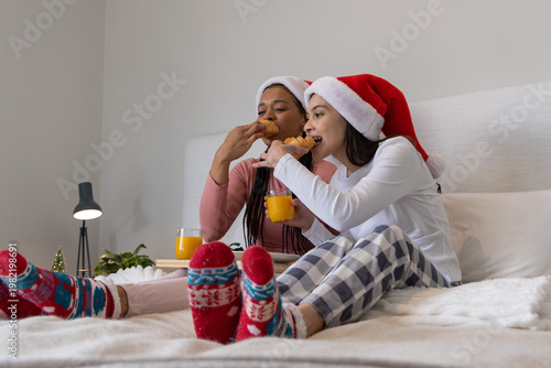 Two adult females sitting on bed wearing Santa hats and pajamas, sharing pastries and orange juice