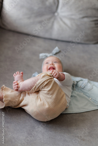 Slika na platnu Joyful baby girl with bow headband playing with her feet while lying on a sofa
