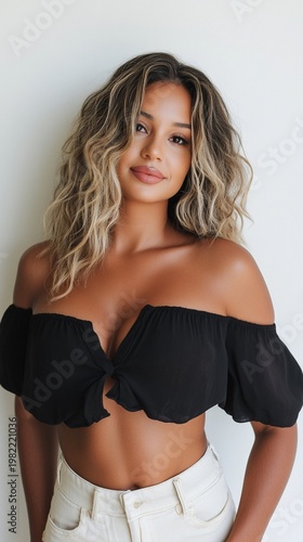 Beautiful young woman with wavy blonde highlighted hair wearing black off-shoulder crop top and white high-waisted pants against light background, showcasing modern fashion style.