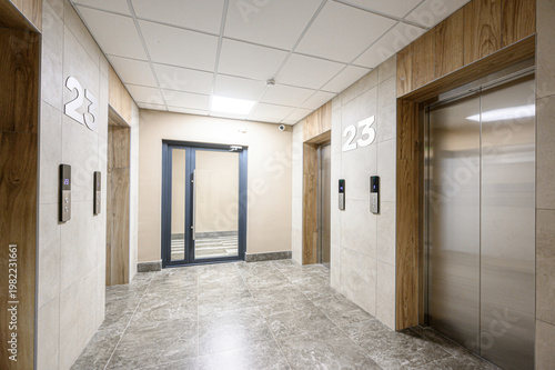 Photos A modern elevator lobby with light beige tiled walls, wooden accents, stainless steel elevator doors, gray marble flooring, recessed ceiling lights, numbered panels (23), glass entrance door