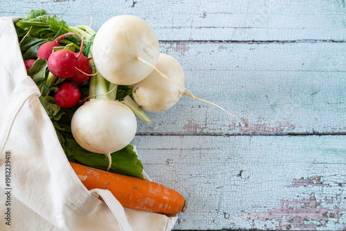 A reusable cotton shopping bag lies on a rustic table with fresh vegetables spilling out, top view.