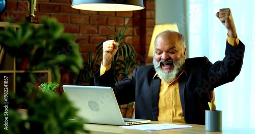 Indian senior businessman or corporate executive showing a successful gesture, such as a fist pump or confident smile, while working on his laptop at a desk in a cozy office with moody lighting.
