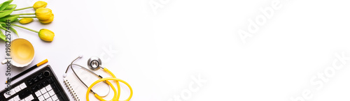 Long wide banner with doctor office desktop with keyboard, cup of tea, medical stethoscope and yellow tulips on white background, copy space for your design. Greeting card for medics template.