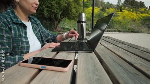 Woman Engages in Remote Work Outdoors, Typing on Laptop at a Picnic Table in Nature