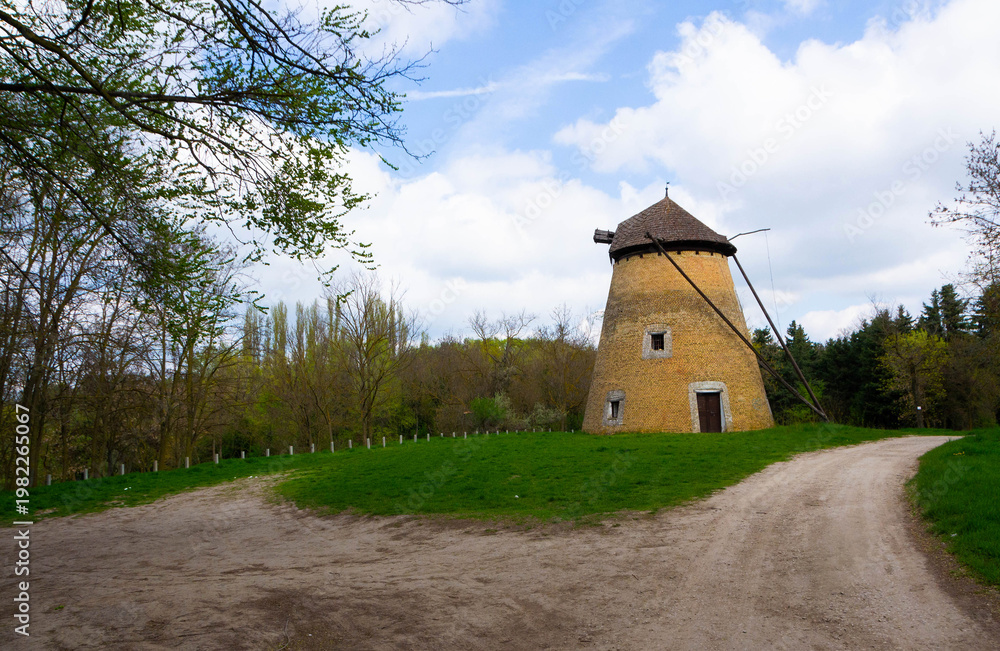 Fototapeta premium Old traditional windmill without sails on a green hill with a dirt road and trees under blue sky with clouds. Rustic countryside landscape, historical architecture, travel and rural concept.