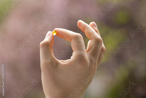 Closeup of a person holding a vitamin D3 capsule with flowers in the background. Concept of supplementing vitamin D in spring.