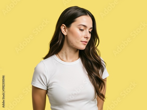 Young woman looking down thoughtfully against a yellow background. Casual female portrait in a white t-shirt in studio