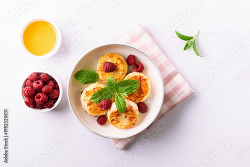 Cottage cheese pancakes with honey, raspberry and mint leaves on white ceramic plate on white marble table top view. Healthy homemade dessert.