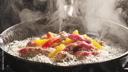 Slow motion close-up of sizzling hot peppers flaming in cast iron skillet on dark background