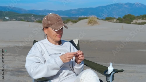 Woman knitting outdoors on a sandy beach chair. Middle-aged woman in a cap relaxing in a folding chair on a sandy beach, enjoying her knitting hobby while surrounded by a tranquil landscape of dunes